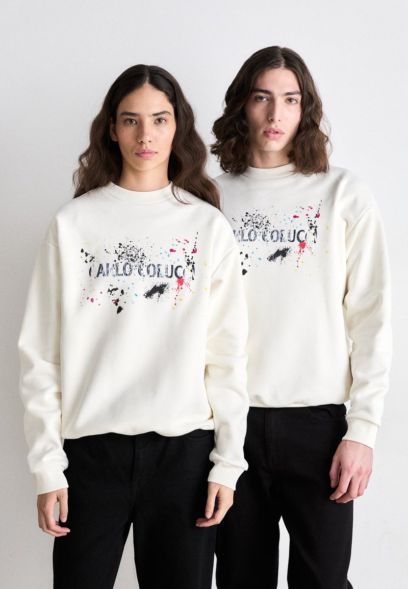 Sweat-shirts crème avec un imprimé graphique comportant des éclaboussures de couleur et du texte, portés avec un pantalon noir ; coupe décontractée, poignets côtelés.