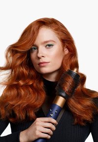 Uno strumento per acconciare i capelli, arrotondato, con una testa di setole nere e dettagli dorati. Il manico è blu e presenta dei tasti di controllo, ed è impugnato da una persona con capelli rossi.