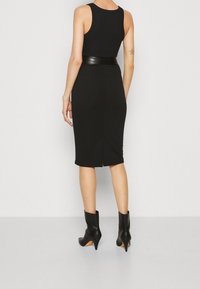 Černé přiléhavé šaty bez rukávů, s páskem v oblasti pasu, délka ke kolenům a rozparek na zadní straně. Spárováno s černými kotníkovými botami. Hladká textura.