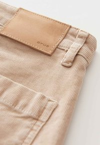 Pantalon en velours côtelé beige avec une surface texturée. Comprend un patch rectangulaire en cuir cousu portant la mention « OPUS » sur la ceinture.