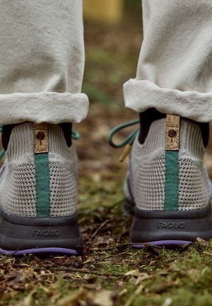 Baskets en maille gris clair avec semelles noires et boucles arrière turquoise, portées avec un pantalon beige retroussé, debout sur le sol de la forêt.