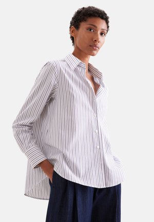 Vrouw draagt een losse witte blouse met dunne verticale zwarte strepen en donkerblauwe broek, staand met handen in de zakken.