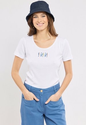 Femme souriante, portant une chemise blanche à manches courtes avec un petit motif de poissons multicolores, un pantalon bleu et un chapeau seau bleu foncé.