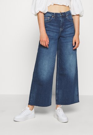 Nainen, jolla on korkeavyötäröiset, leveälahkeiset siniset farkut, joissa on rispaantuneet reunat, ja valkoiset tennarit, yhdistettynä lyhytvalkoiseen, puffihihalliseen toppiin.