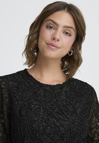 Crna čipkana bluzica s složenim cvjetnim uzorcima, okruglim dekolteom i teksturiranom tkaninom. Ima blago napuhane rukave i delikatnu završnicu.