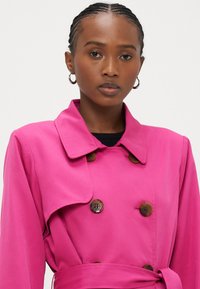 Ροζ καμπαρντίνα με διπλό πέτο, μεγάλο γιακά και καφέ κουμπιά. Διαθέτει ζώνη στη μέση και απαλή υφή υφάσματος.