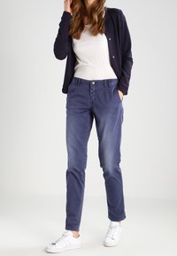 Vrouw in een marineblauwe broek, witte sneakers, een witte top en een marineblauwe knopen cardigan, staand met een hand in de zak.