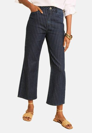 Kvinde iført marineblå, bredbenede, cropped bukser med striber, beige vævede sandaler, hvid bluse og guld armbånd på håndleddet.