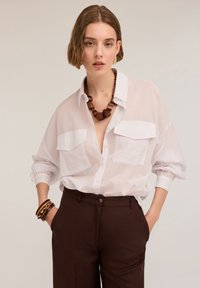 Cămașă albă cu butoni, cu două buzunare frontale și mâneci răsfrânte. Pantaloni maro cu talie înaltă. Accesorizată cu un colier mare din lemn.
