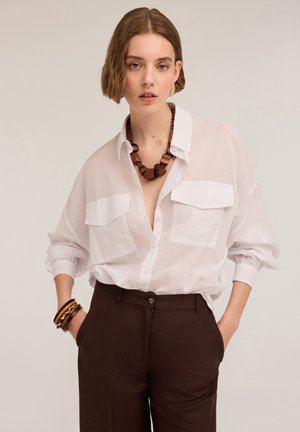 Weißes Hemd mit Knopfleiste und zwei vorderen Taschen, hochgekrempelte Ärmel. Braune, hoch sitzende Hose. Mit einer großen Holz-Halskette accessorized.