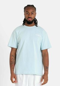 Camiseta de algodón azul claro con cuello redondo y mangas cortas. Presenta un pequeño logo blanco en el lado izquierdo del pecho. Textura suave.