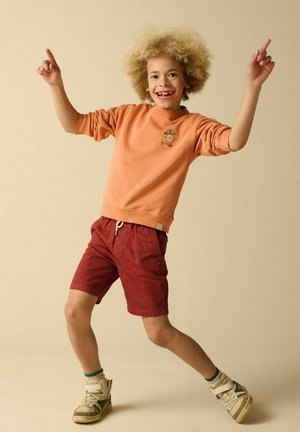 Enfant aux cheveux bouclés portant un sweat-shirt orange et un short rouge, souriant et pointant vers le haut avec les deux mains tout en se tenant sur un pied.