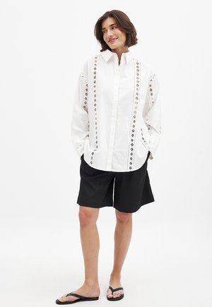 Weißes Baumwollhemd mit aufwendigen Ausschnittdetails an der Vorderseite, kombiniert mit schwarzen Shorts und Flip-Flops, vor einem weißen Hintergrund stehend.