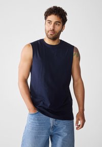 Tengeri kék ujjatlan póló, pamutból készült, kerek nyakkal, sima textúrával, laza szabással és minimalista dizájnnal, világos farmer sorttal párosítva.