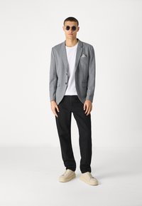 Grå kavaj med notchade laplar, svarta byxor, vit t-shirt och kraftiga beige sneakers. Rundade solglasögon och mönstrad fickrekt.