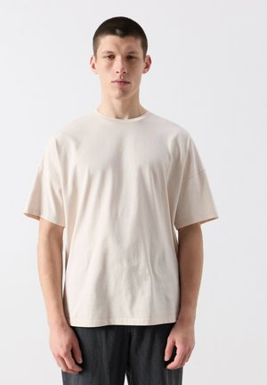 Noor mees lühikeste tumedate juustega, kandes üle suure suurusega beeži lühikese varrukaga T-särki ja tumedaid pükse, seisab ühevärvilise heleda tausta ees.