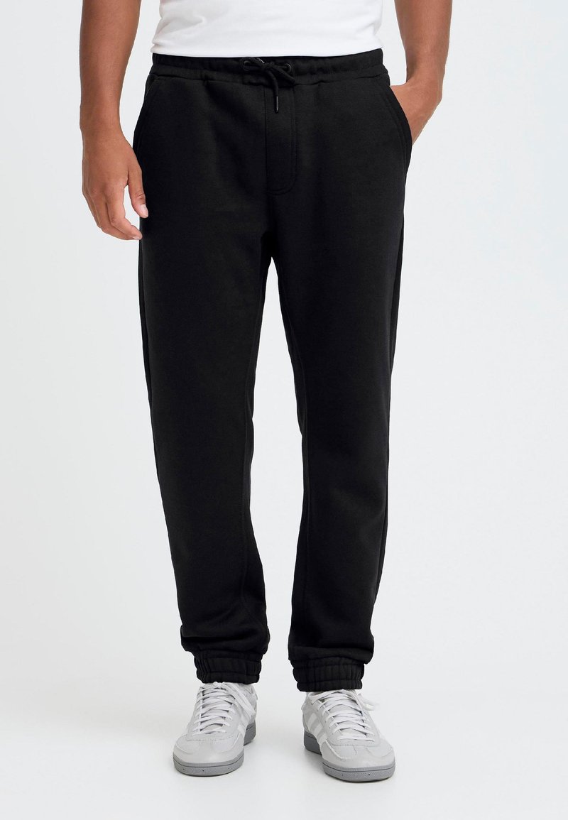 Bărbat purtând pantaloni jogger negri cu manșete elastice, adidași albi și un tricou alb, cu mâinile în buzunare, stând pe un fundal simplu.