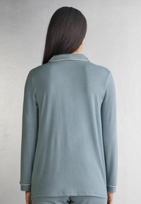 Lichtblauwe lange mouw top met een subtiele kraag en gouden piping accenten. Soepele textuur en relaxte pasvorm, achteraanzicht getoond.