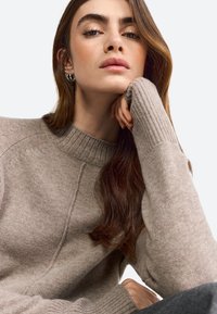 Maglione beige a maglia con colletto alto a coste e polsini a coste. Il modello poggia il mento sulla mano, mostrando una pelle liscia e capelli lunghi e mossi.