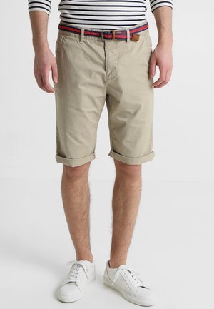 Homme portant un short beige retroussé avec une ceinture tissée rouge et bleue, des baskets blanches et une chemise à rayures à manches longues.