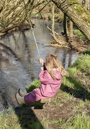 Kind in pinkem Regenanzug sitzt auf einer Holzschaukel, hält ein grünes Seil, neben einem Bach mit Grünflächen und Bäumen im Hintergrund.