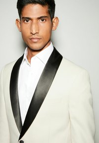 Blazer de couleur crème avec un revers en satin noir, doté d'une poche unique à gauche et d'une chemise blanche en dessous, présenté sur un fond gris.