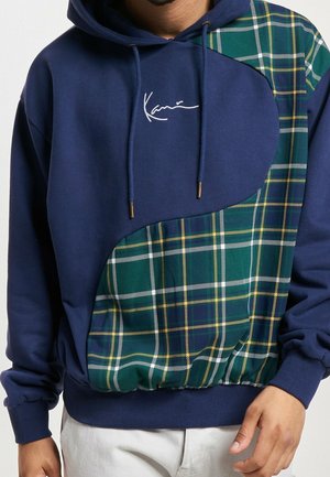 Marineblauer Kapuzenpullover mit grün-gelbem Kariereinsatz auf der Vorderseite, Kordeln mit Metallenden und weißer, gestickter Signatur auf der Brust.