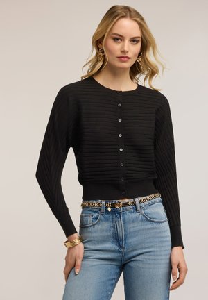 Lyshåret kvinde iført sort ribbet cardigan med knapper, blå højtaljede jeans med flettet bælte, gyldne øreringe og armbånd mod ensfarvet baggrund.