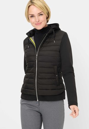 Frau trägt eine schwarze, gesteppte Kapuzenweste über einem schwarzen Langarmshirt und graue Hahnentrittmuster-Hosen, lächelnd mit leicht erhobener Hand.