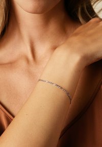 Silverkedja armband på en handled, med länkade ovala och cirkulära former, slät textur och polerad yta mot varmt tonat tyg.