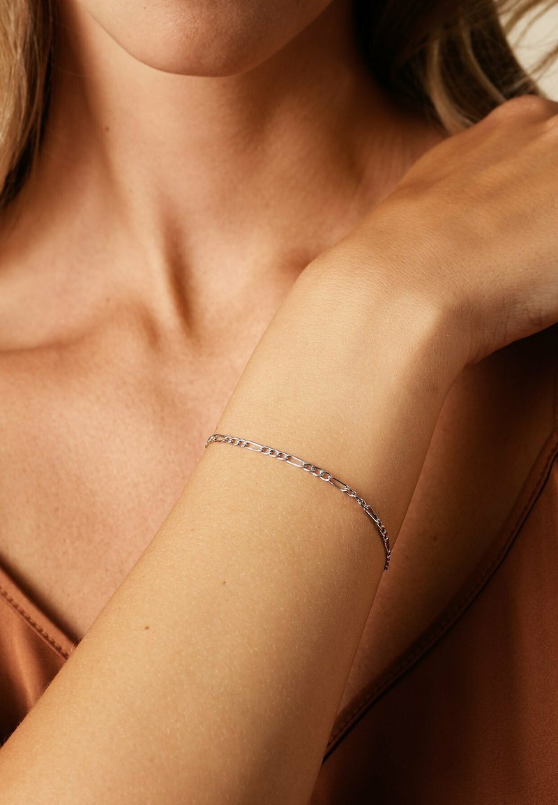 Silverkedja armband på en handled, med länkade ovala och cirkulära former, slät textur och polerad yta mot varmt tonat tyg.