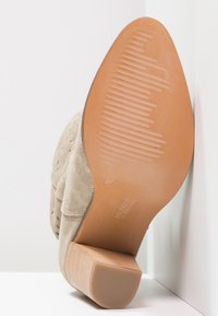 Botín de ante beige hasta el tobillo con un tacón bajo y cuadrado; parte superior de cuero texturizado con perforaciones; suela de goma marrón claro con detalles estriados.
