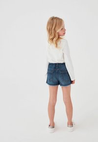 Jeansshorts med fransad nederkant, visad med en vit långärmad skjorta. Har en bakre midjeband och två bakfickor, tillsammans med vita sneakers.
