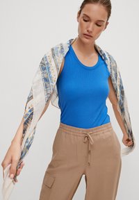 Top a maniche lunghe blu con righe e pantaloni beige con vita elastica. Una sciarpa leggera e decorata pende dalle spalle, con accenti blu e oro.