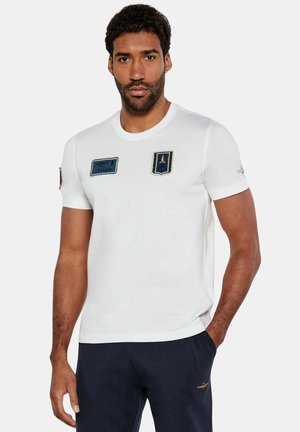 T-shirt bianca a maniche corte con scollo tondo, caratterizzata da tre toppe ricamate in blu e nero. Tessuto morbido, vestibilità comoda.
