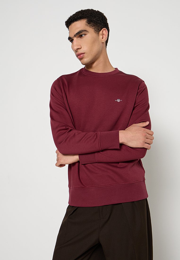 Joven con cabello corto y rizado, vistiendo una sudadera de manga larga burdeos y pantalones oscuros, de pie con los brazos cruzados, mirando hacia la izquierda.