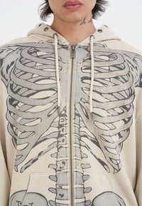 Felpa con zipper beige caratterizzata da un design grafico scheletrico in grigio. Ha un cappuccio con lacci e tasche frontali con polsini a coste.