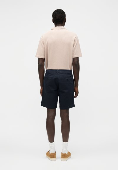 Homme portant un polo manches courtes beige clair, un short bleu marine foncé avec poches arrière, des chaussettes blanches et des baskets en daim beige, de dos sur fond blanc.