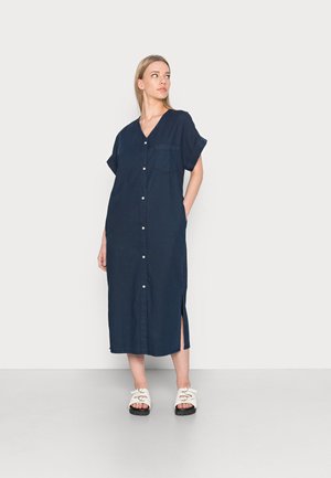 Vestido botão azul marinho com mangas curtas, colarinho plano, bolso frontal, fendas laterais, feito de tecido leve. Combinado com sandálias brancas.