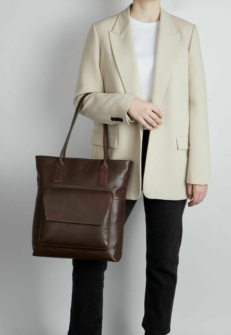 Personne portant un blazer beige et une chemise blanche tient un grand sac cabas en cuir marron foncé avec poche avant, debout devant un fond uni.