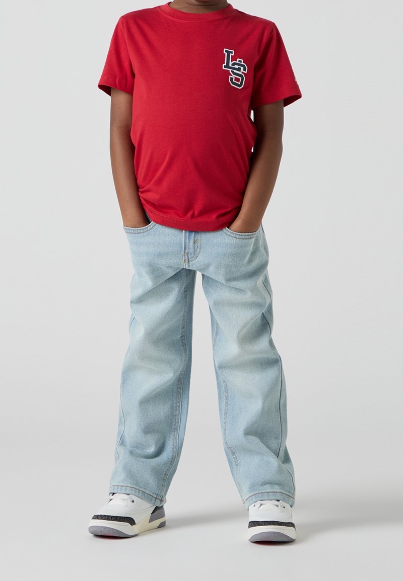 Camiseta de algodón roja con un logo estampado, combinada con pantalones vaqueros de mezclilla azul claro, con un ajuste relajado y dos bolsillos delanteros. Zapatillas blancas completan el conjunto.