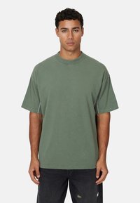 T-shirt verde in cotone oversize con colletto rotondo, maniche corte e cuciture a contrasto. Abbinata a jeans scuri strappati.