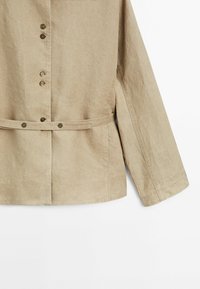 Vaaleanbeige pellavakangasta takki, jossa on pitkät hihat, nappikiinnitys edessä ja vyötärö vyöllä. Takissa on sileä rakenne ja minimalistinen muotoilu.