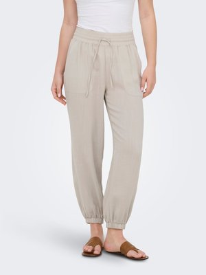 Femme portant un pantalon beige avec cordon de serrage, ourlet élastique, poches avant et sandales plates marron à bouts ouverts, debout sur un fond uni.