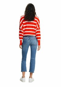 Femme aux cheveux bouclés portant un pull rayé rouge et blanc, un jean court bleu et des baskets blanches, debout, de dos sur fond blanc.