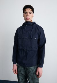 Navy blue pullover jacket in lightweight fabric with a high collar, drawstring waist, and multiple front pockets; subtle texture visible.