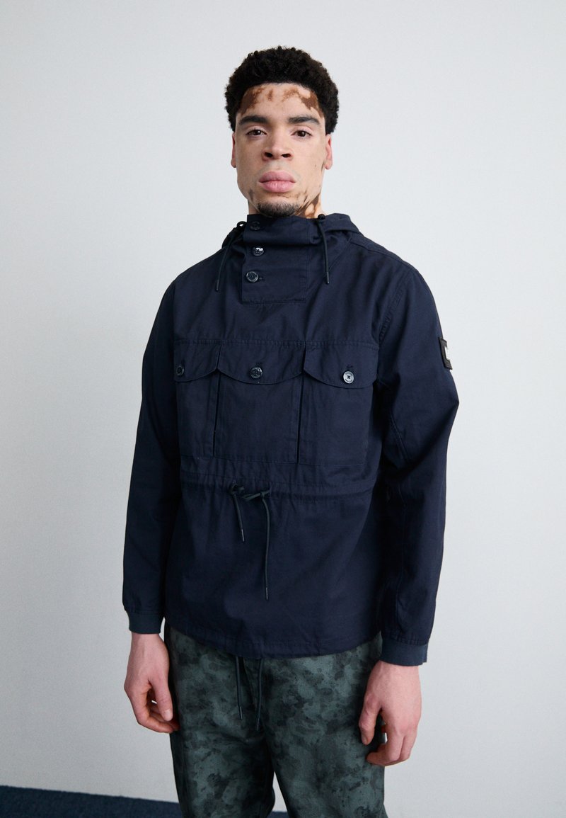 Navy blue pullover jacket in lightweight fabric with a high collar, drawstring waist, and multiple front pockets; subtle texture visible.