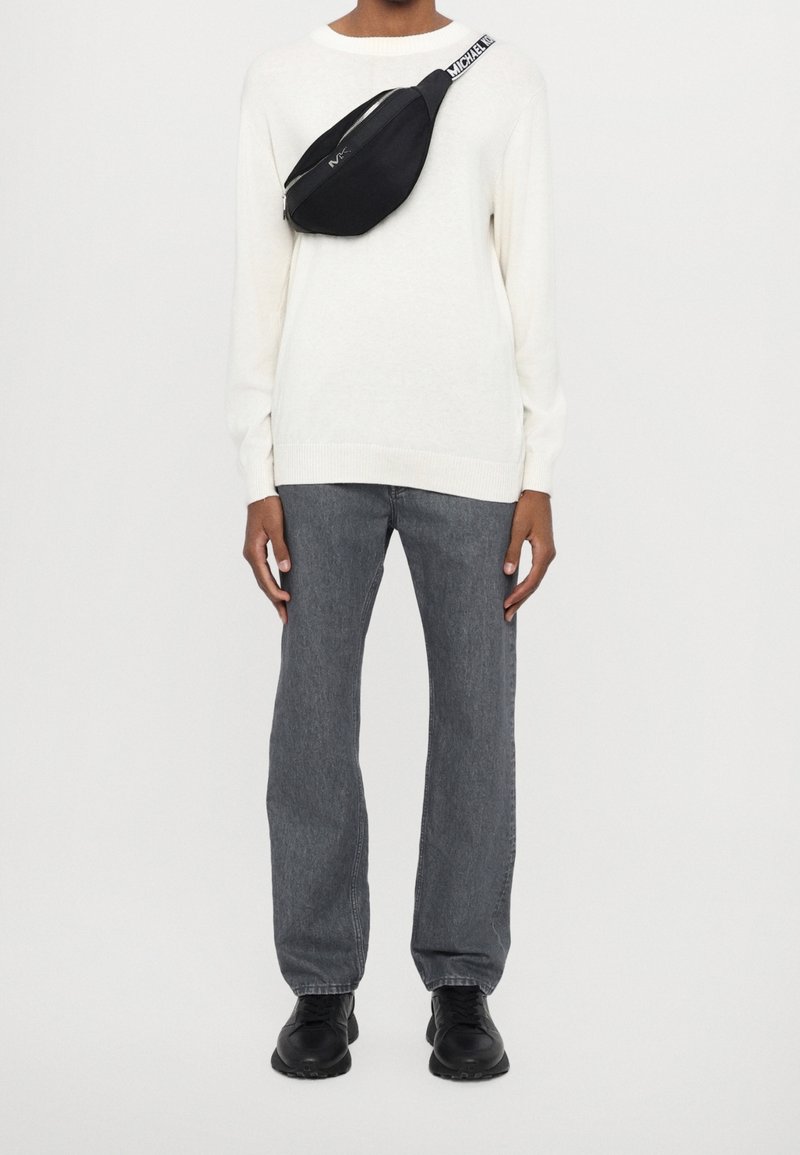 Personne portant un pull blanc, un jean gris droit, des chaussures noires et un sac bandoulière noir avec une sangle de marque.