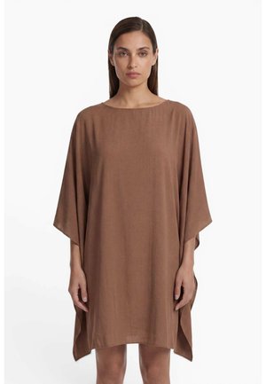 Femme aux cheveux bruns et lisses portant une robe marron ample jusqu'aux genoux avec de larges manches, debout devant un fond blanc uni.
