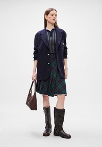 Ναυτικό σακάκι με χρυσά κουμπιά, ριγέ πλεκτό πόλο, καρό πράσινη φούστα, καφέ δερμάτινες μπότες μέχρι το γόνατο και καφέ τσάντα. 